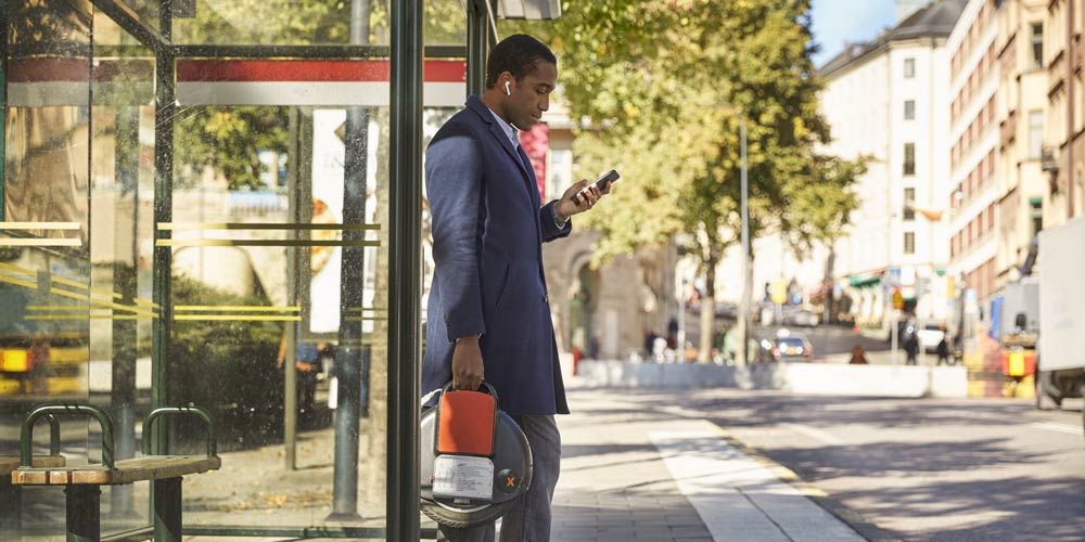 Em man med airpods och som tittar i sin telefon står vid en busshållsplats i Stockholm.