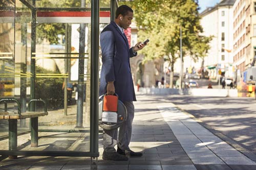 En man med airpods och som tittar i sin telefon står vid en busshållsplats i Stockholm.